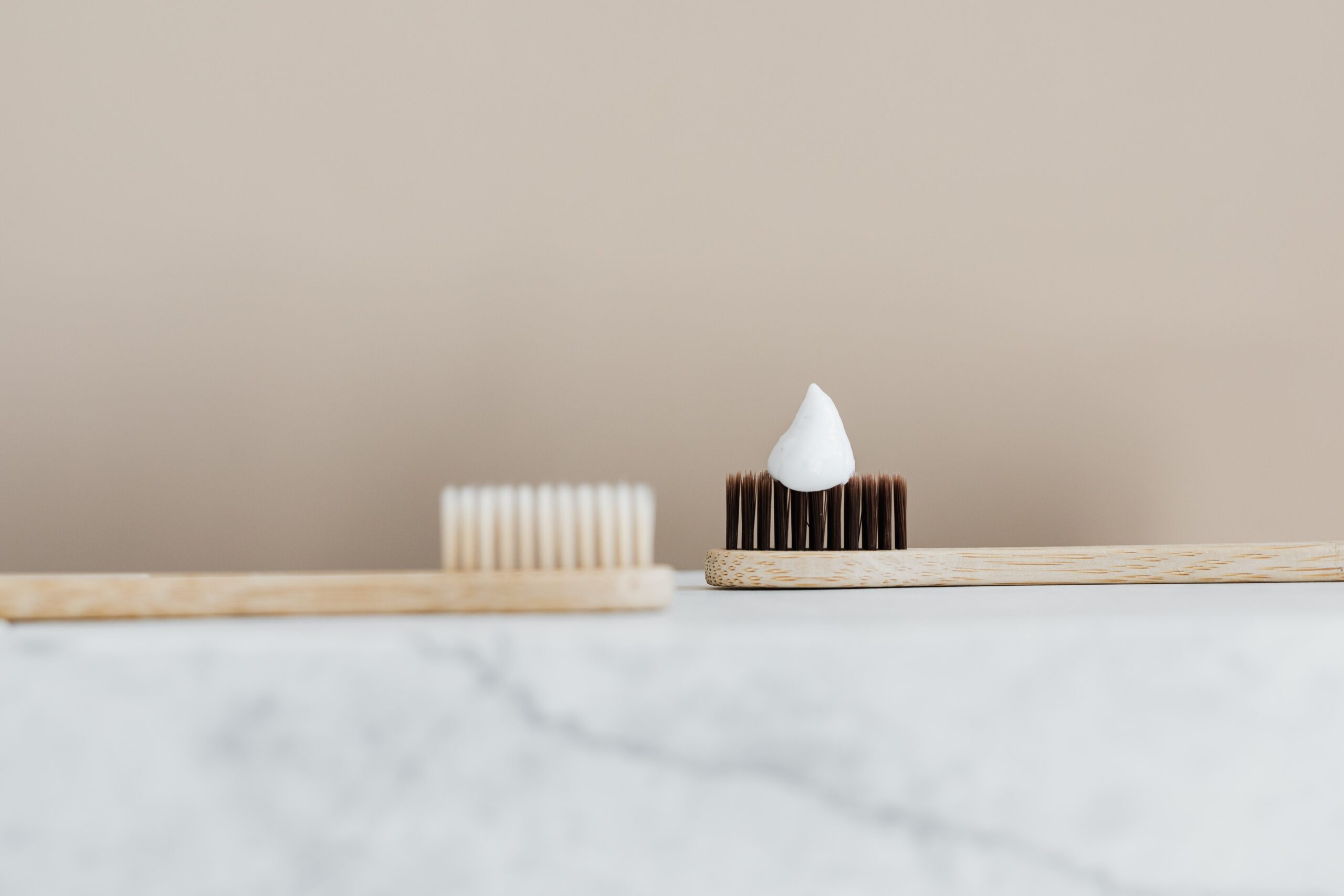 Two wooden toothbrushes on a marble edge one with white bristles and one with black bristles and a dollop of toothpaste.